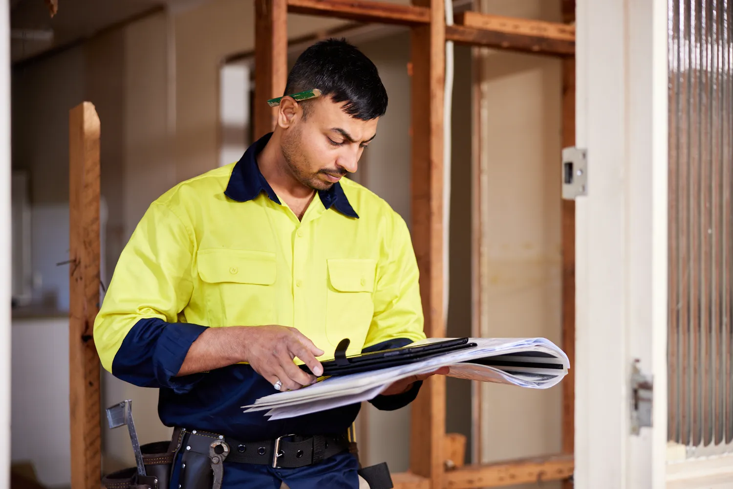 A builder wearing hi-vis clothing standing in a worksite reading paperwork and holding a mobile device