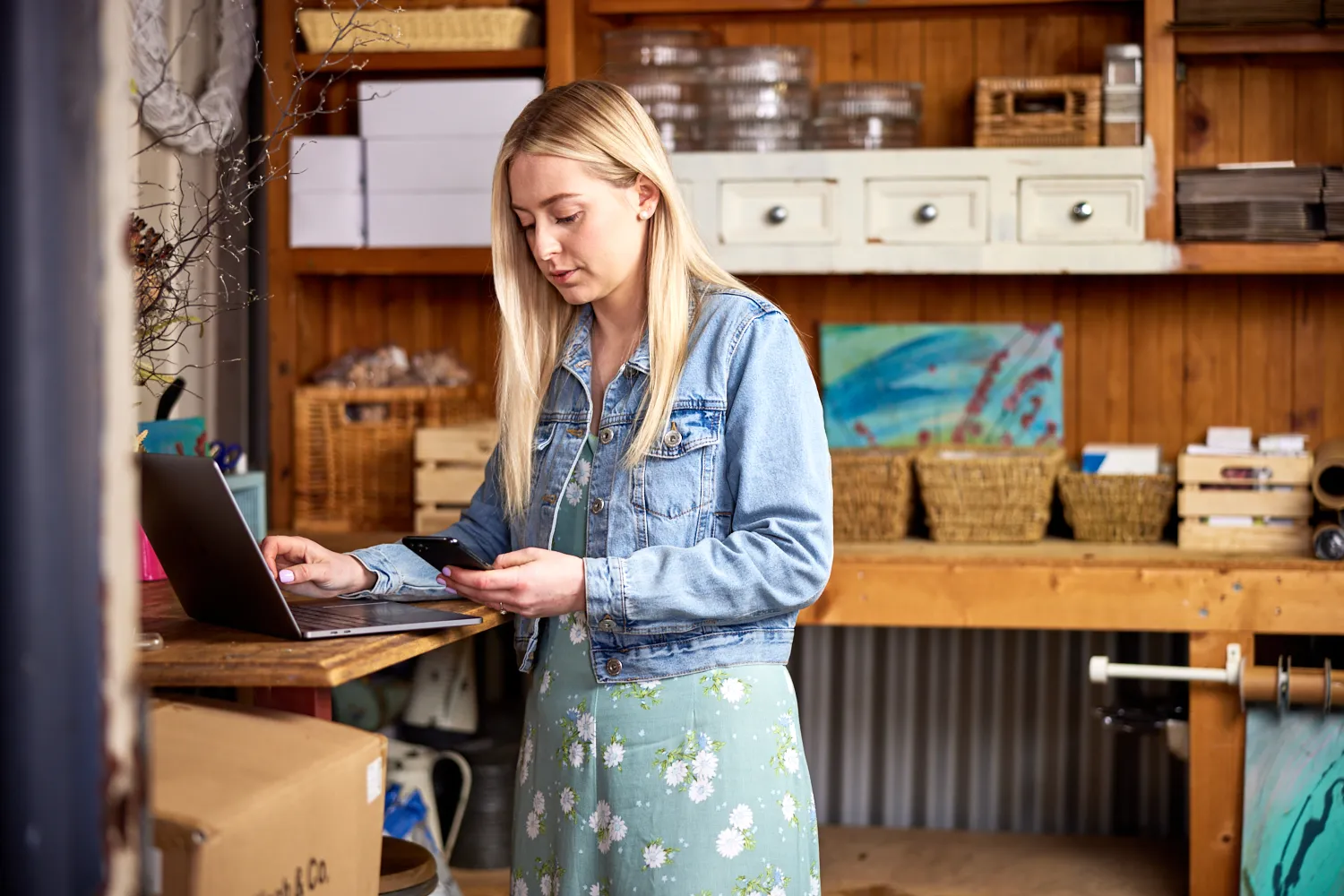 A person looking at a mobile phone, standing at a laptop in a shed surrounded by florist supplies