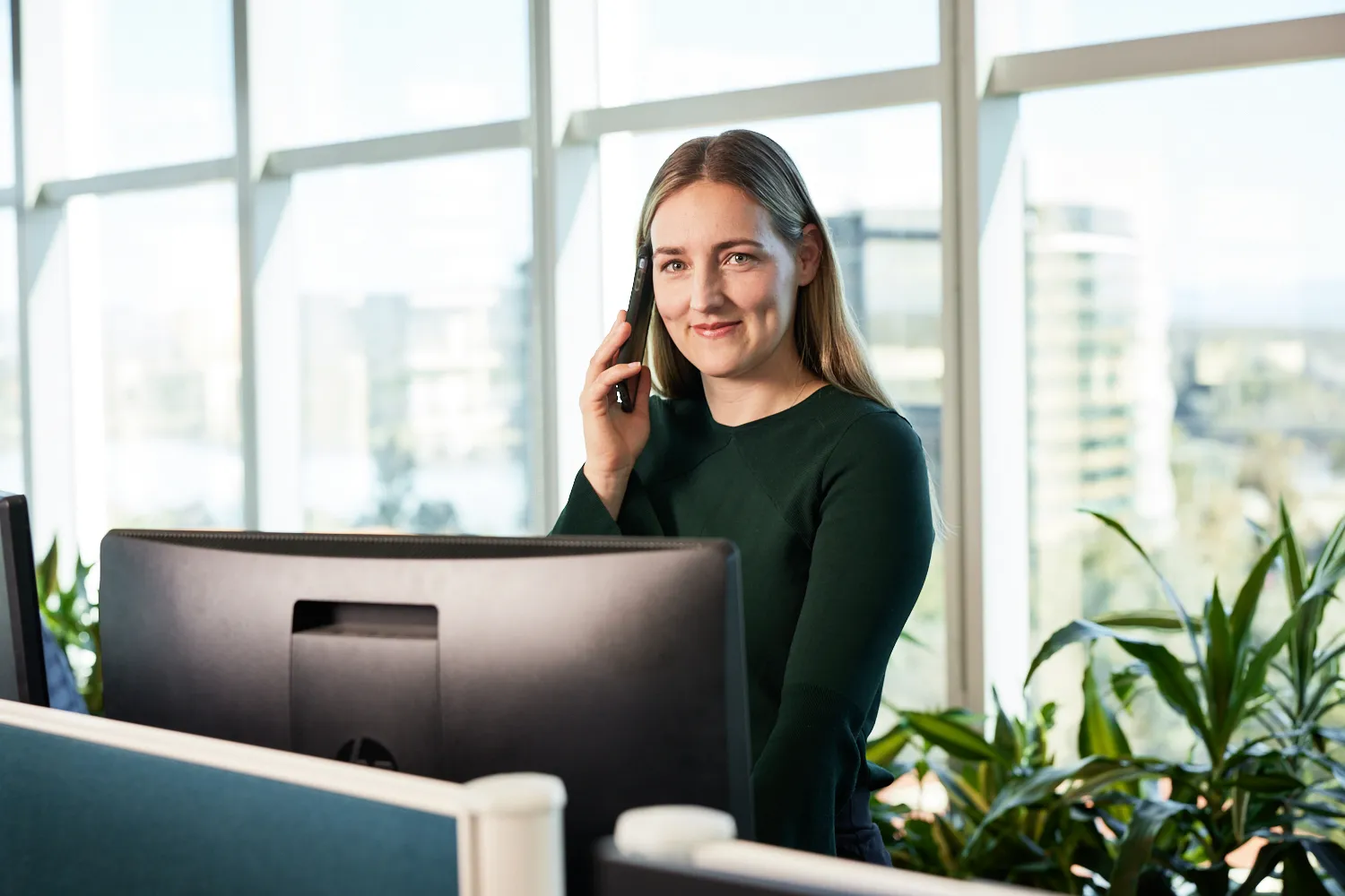 A smiling adult sitting at a computer and holding a phone to their ear